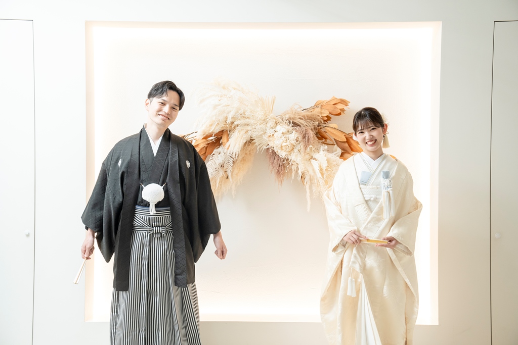 ドライフラワーのアーチを背に並んで立つ、白無垢と黒紋付袴の新郎新婦の和装スタジオフォト。