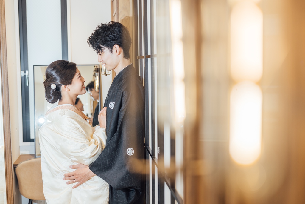 白無垢姿の新婦と黒紋付袴姿の新郎が鏡越しに寄り添う、格子戸の前で見つめ合う横顔