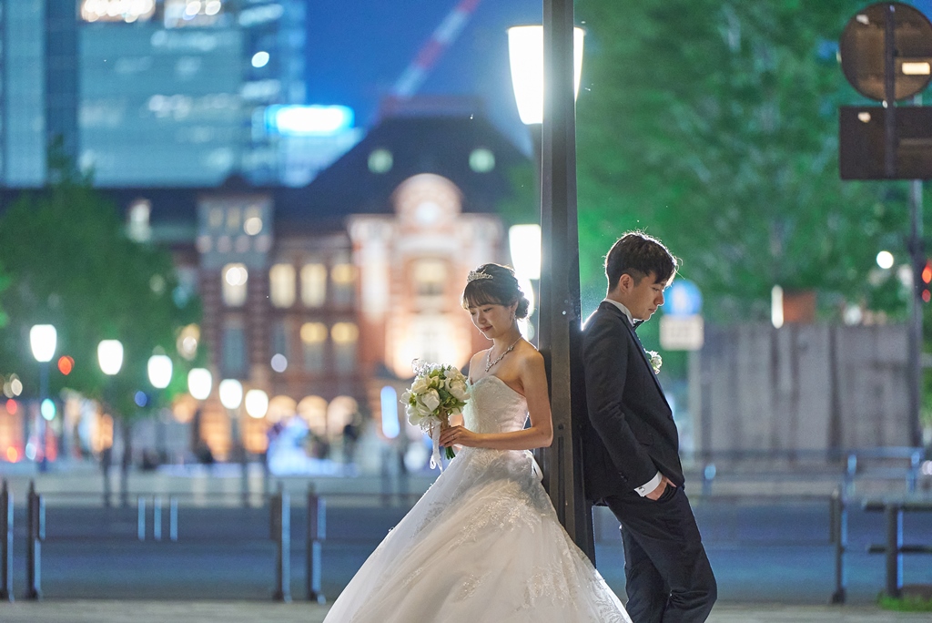 東京駅を背景に、街灯にもたれる新郎とブーケを持つ新婦の夜景ウェディングフォト。