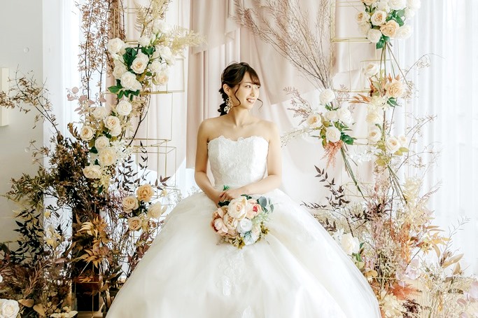 白いストラップレスのウェディングドレス姿の花嫁がドライフラワーのアーチを背景に微笑む、ブーケを手にした立ち姿