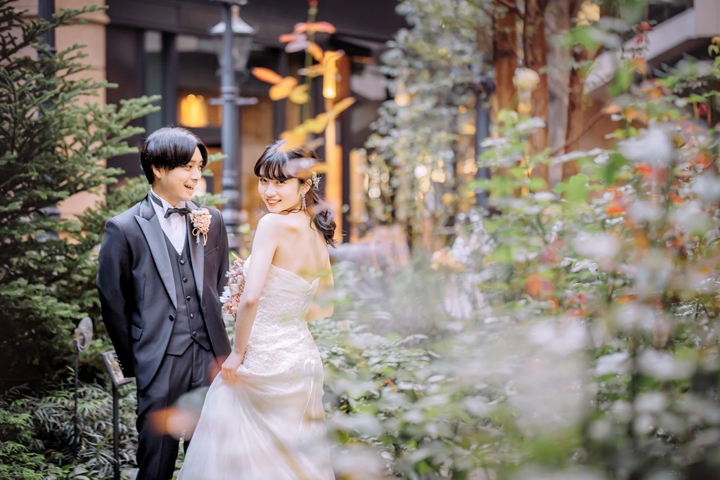 花々に彩られたブリックスクエアの小径で振り返る新婦と寄り添う新郎、レンガ造りの建物を背景にした洋装ロケーションフォト。
