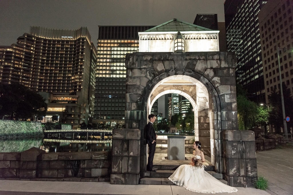 夜景の中、和田倉門の石造りアーチの下で座る新婦とたたずむ新郎、東京の高層ビル群と噴水広場を背景に。