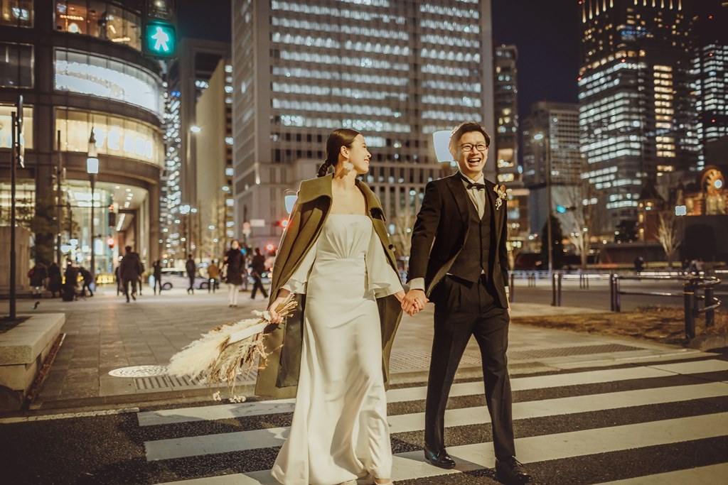 夜の高層ビル群を背景に、横断歩道を手をつないで渡る新郎新婦、新婦はカーキのケープを羽織る。