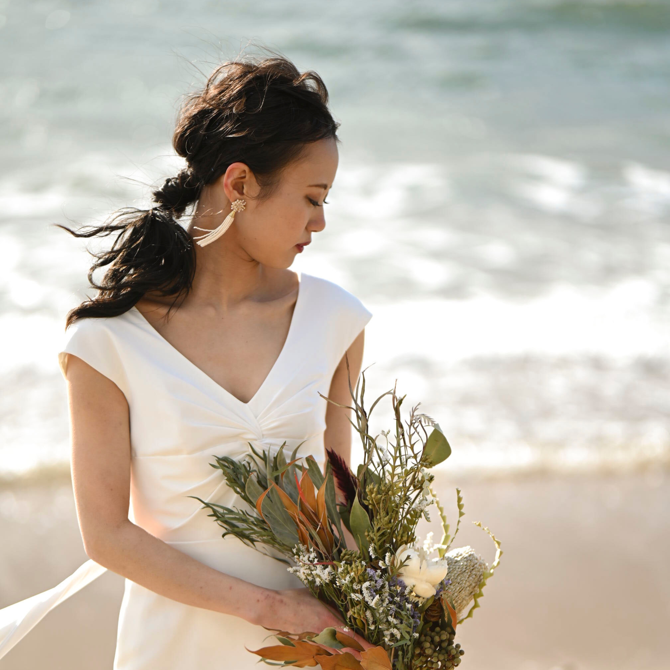 波打ち際で、風になびく髪とドレスの花嫁、ドライフラワーのブーケを持つ横顔。