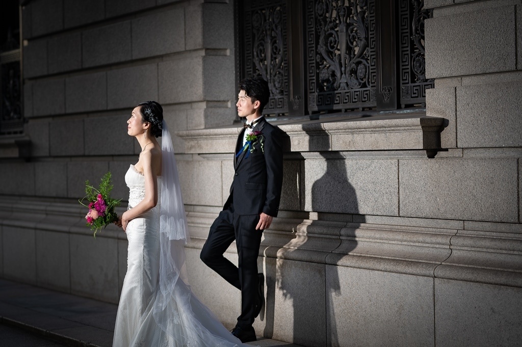 格子装飾窓を背景に、並んで立つヴェール姿の新婦とタキシード姿の新郎。