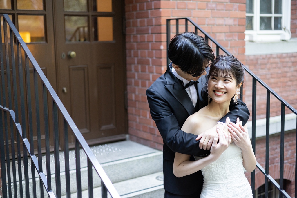 レンガの壁を背景に抱き合う新郎新婦、白いオフショルダードレスの新婦。