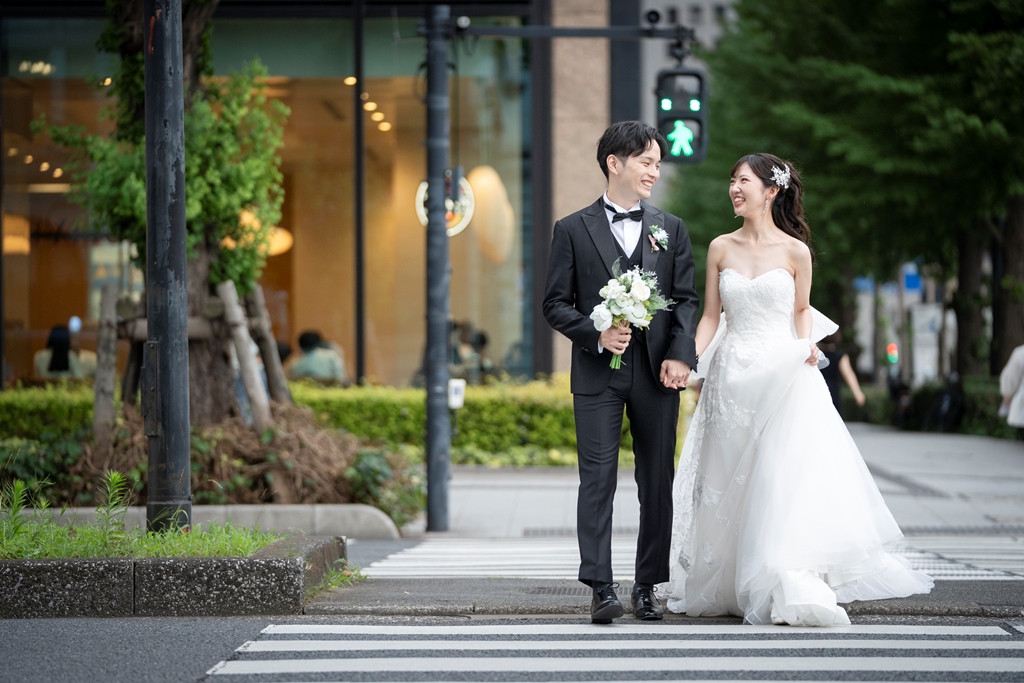 横断歩道を微笑みながら手をつなぎ渡る新郎新婦、白いブーケとタキシード。
