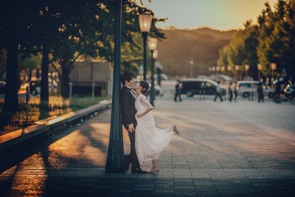 夕暮れの街灯の下、街路樹を背景に足を上げて抱き合う新郎新婦。