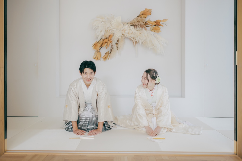 白い羽織袴姿の新郎と白無垢姿の新婦がドライフラワーの壁飾りの前で正座する、結婚写真らしい和のスタジオにて