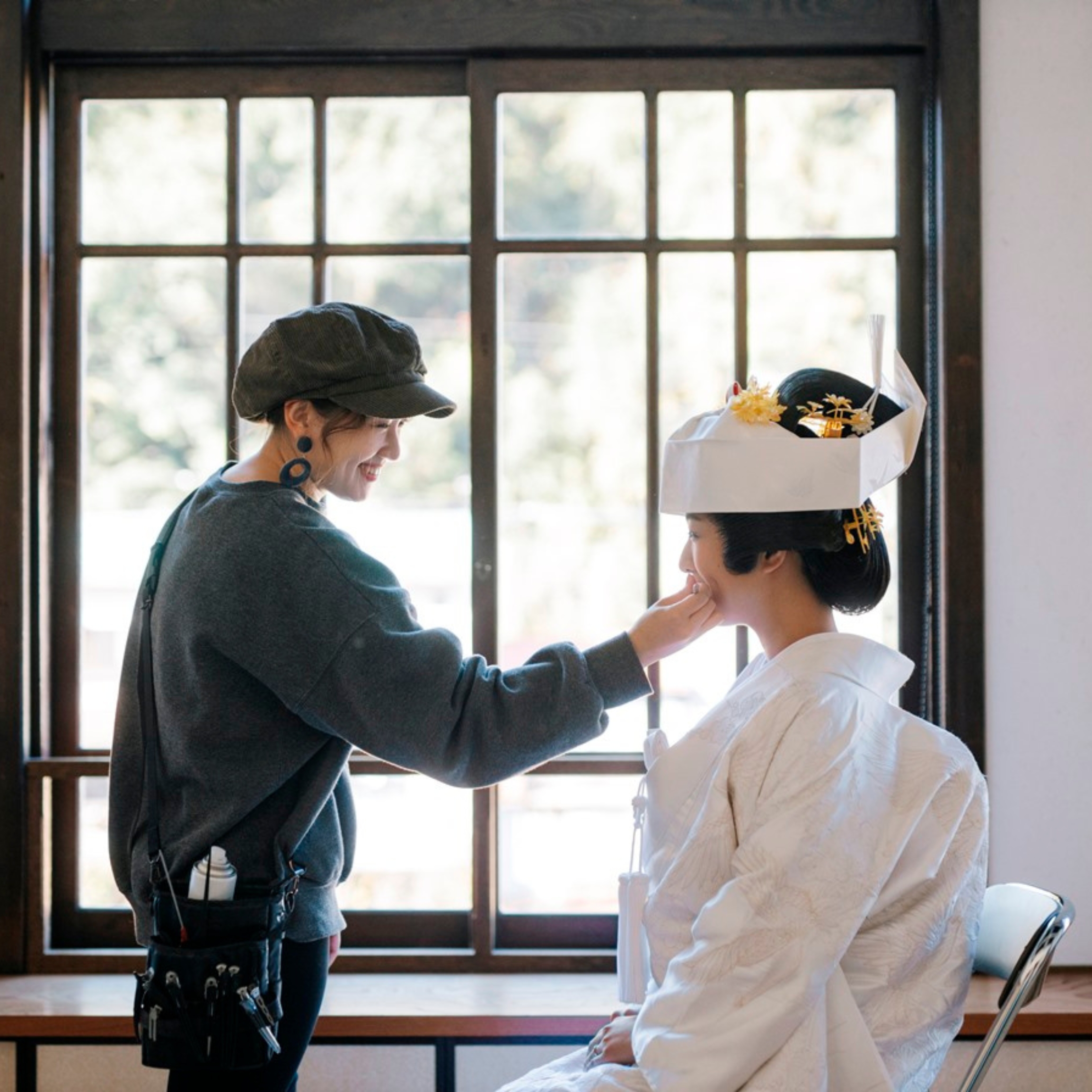 綿帽子を被った白無垢の花嫁の口元に紅を差すヘアメイクスタッフ、窓際での身支度シーン。