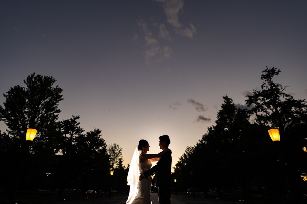 夕暮れ時、街路樹と提灯型の街灯の下、抱き合う新郎新婦のシルエット。