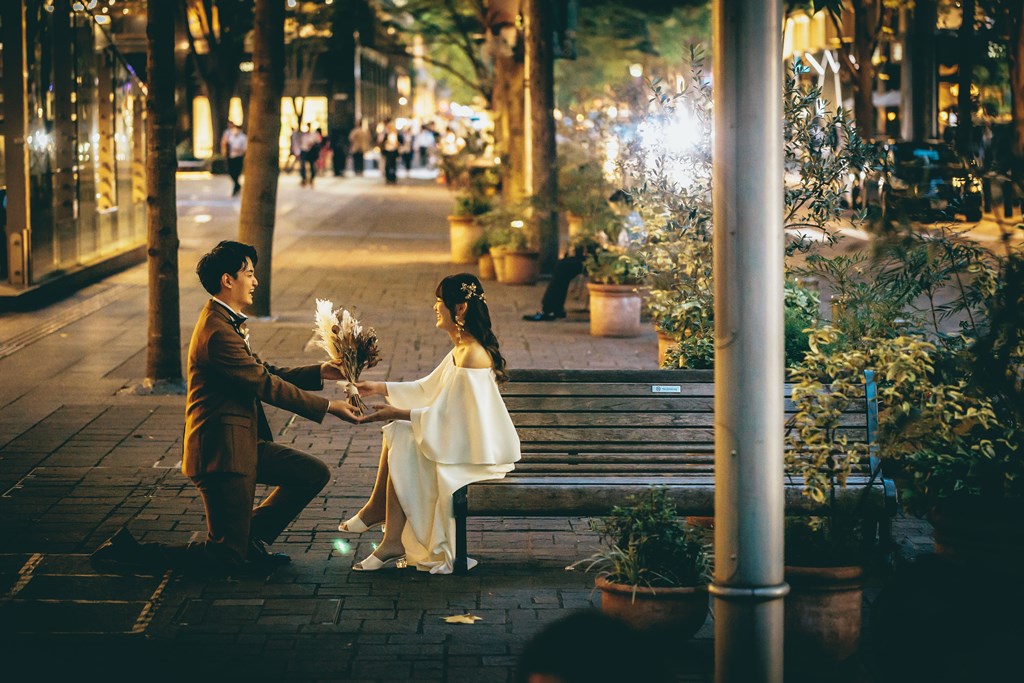 夜の並木道のベンチに座る新婦にドライブーケを差し出す新郎、街の明かりが灯る。