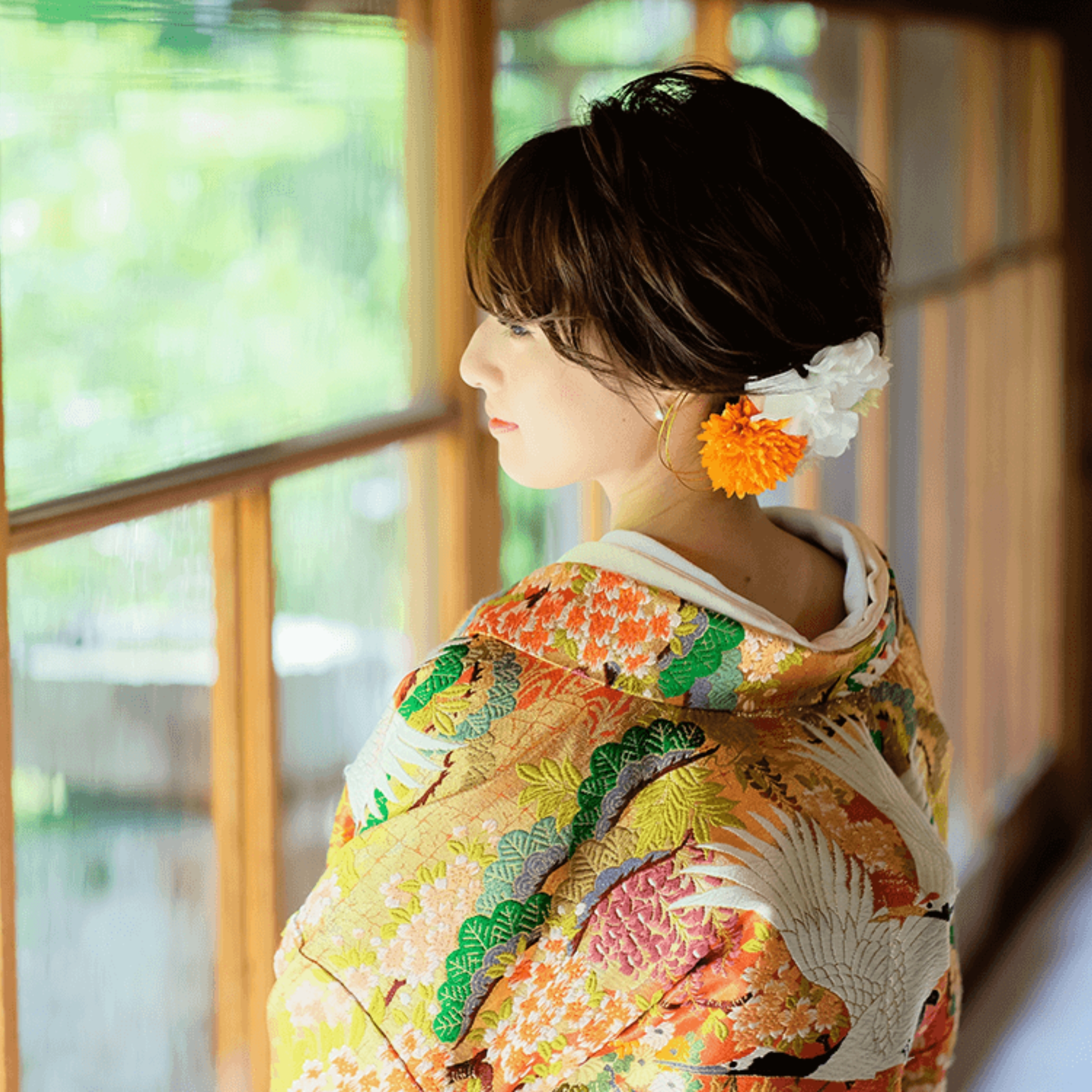 華やかな鶴の刺繍が入った色打掛姿の新婦の後ろ姿、オレンジと白の生花イヤリング、窓辺で外を眺める横顔。