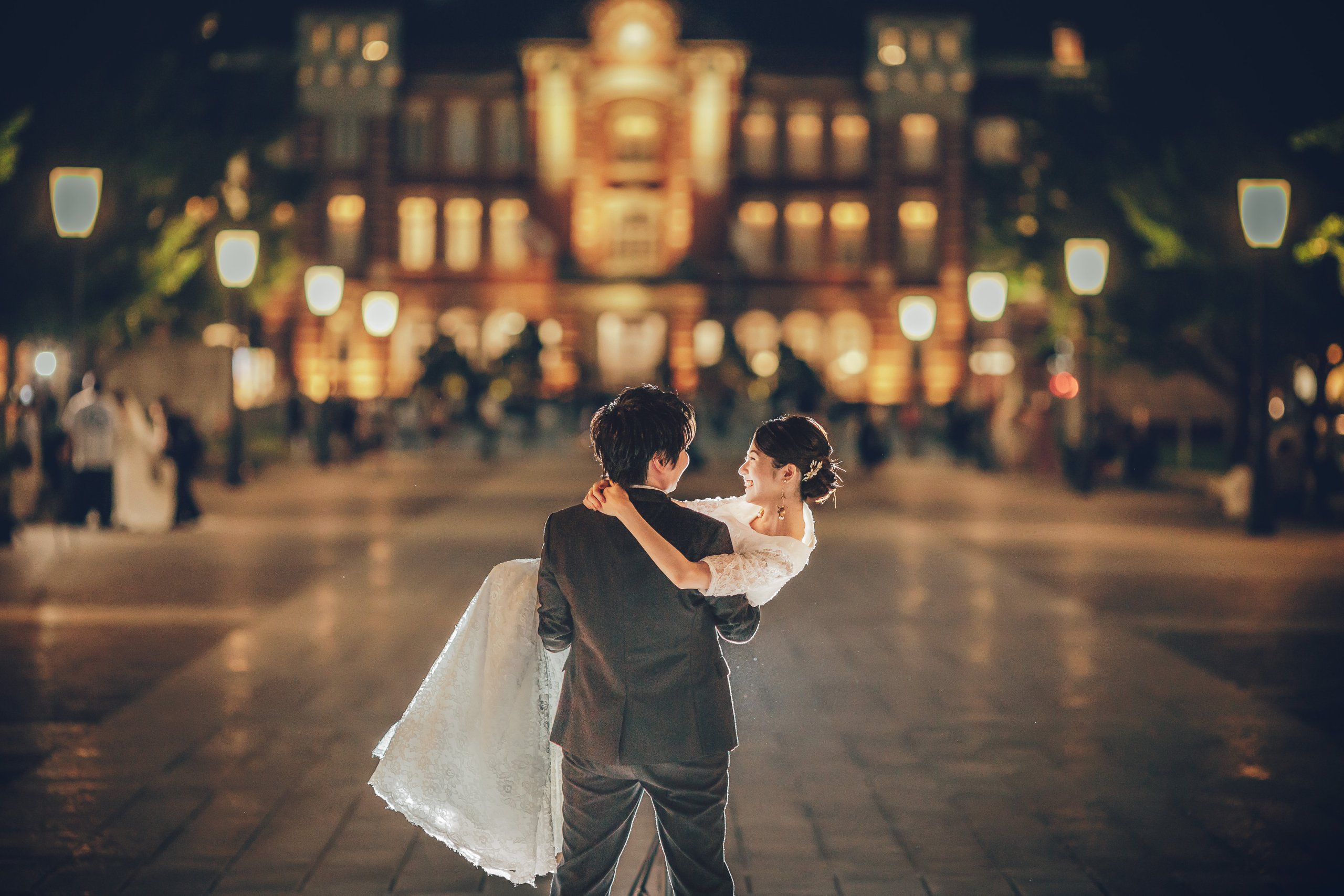 東京駅前の夜景を背景に、新婦を高く抱き上げる新郎のウェディングフォト。