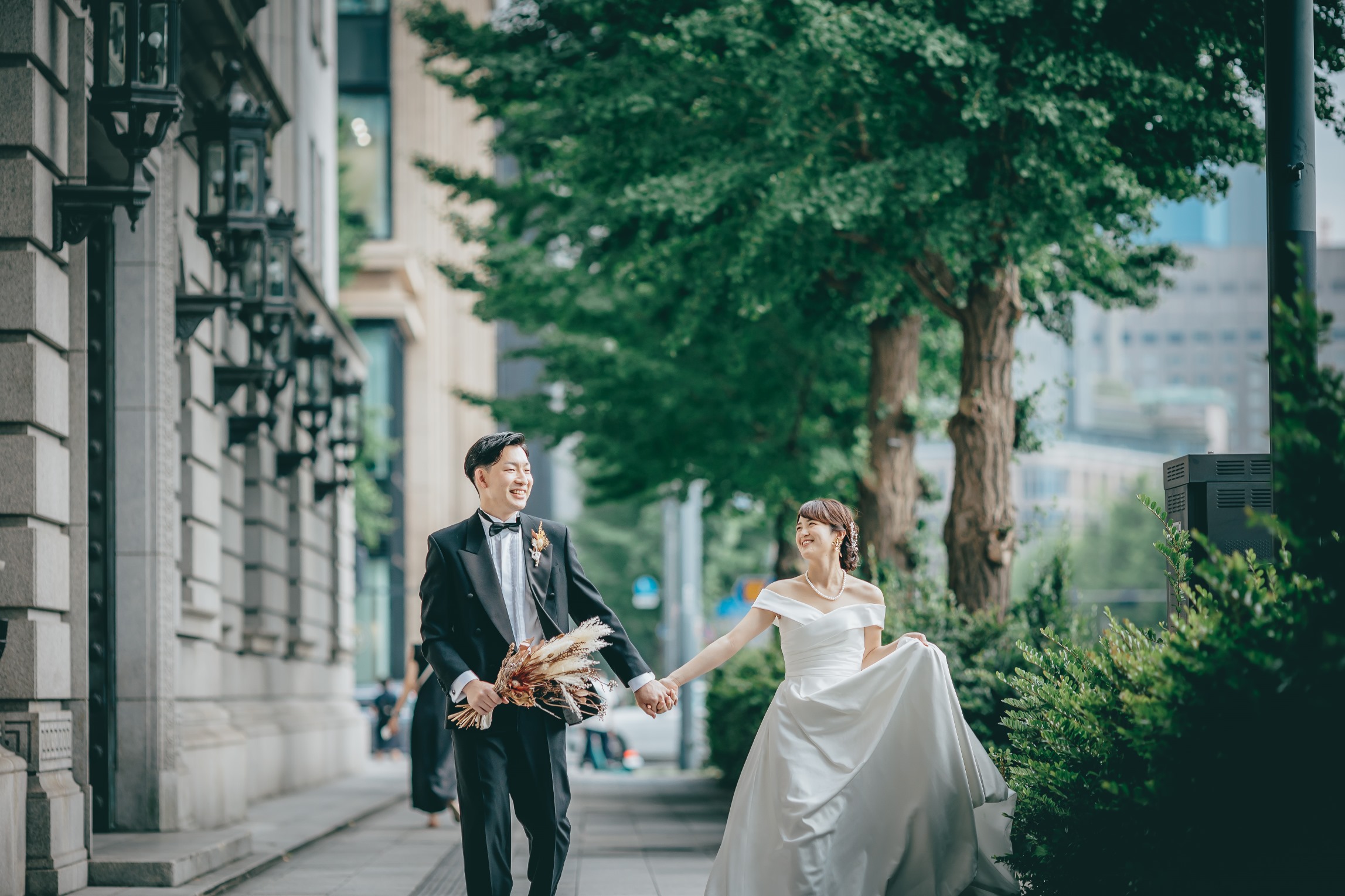 石造りの建物脇の並木道で、ドライブーケを手にした新郎と手をつなぐ新婦。