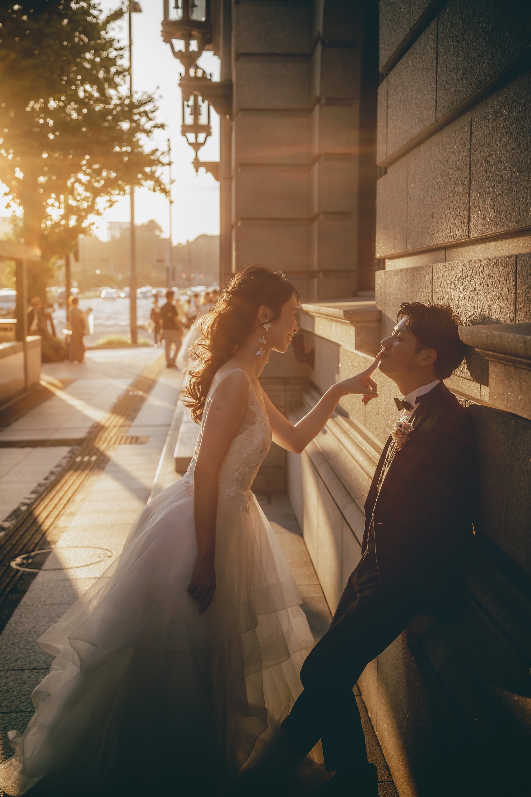 夕陽に照らされた石壁にもたれる新郎、鼻先に指を添える新婦、白いドレスのカジュアルな一枚。