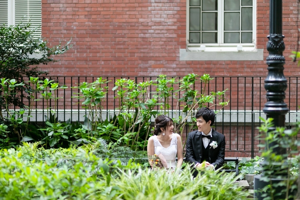 柵越しの植え込みに囲まれたベンチに並んで座る新郎新婦。