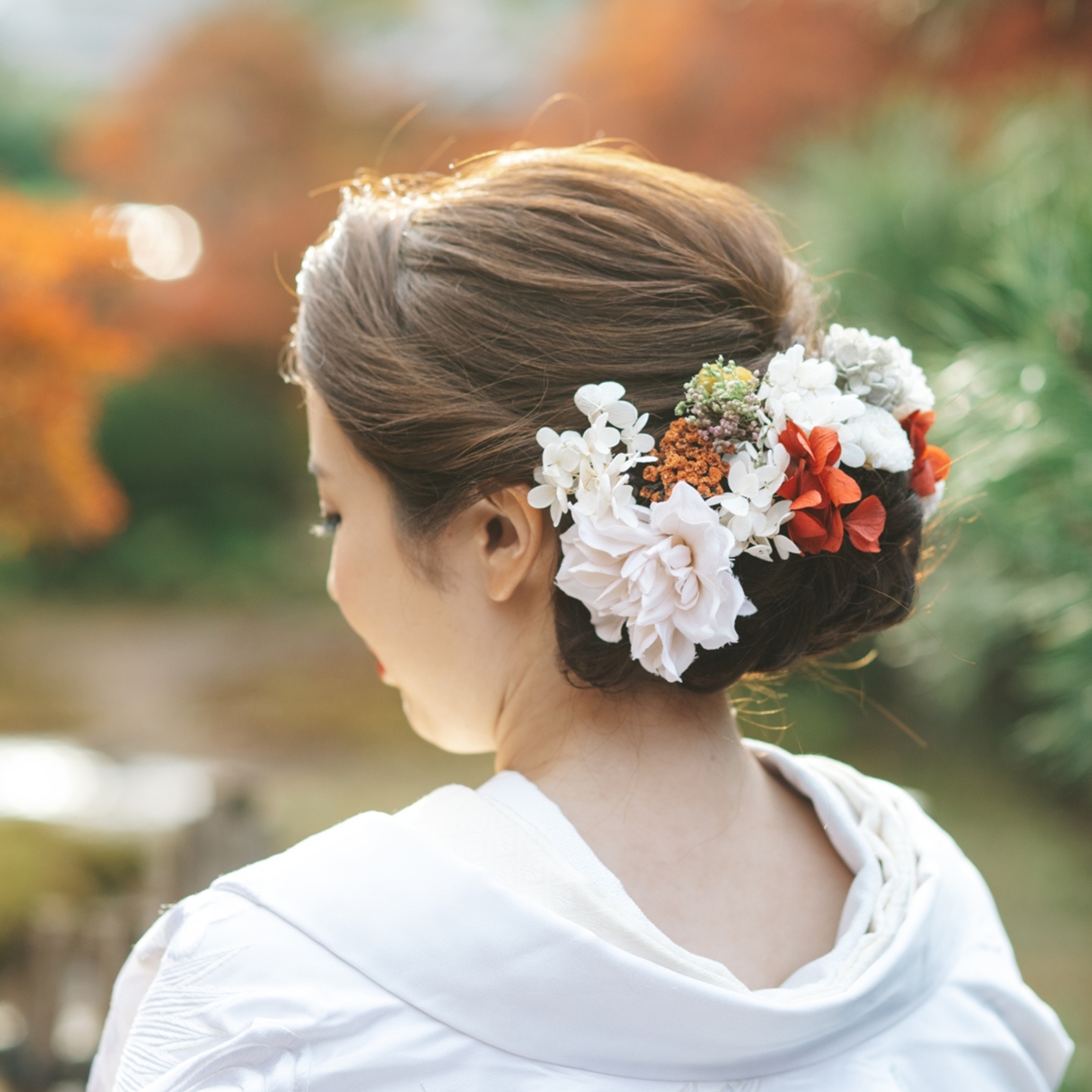 紅葉を背景に、白や赤の生花をあしらった上品な和装ヘアアレンジの後ろ姿。