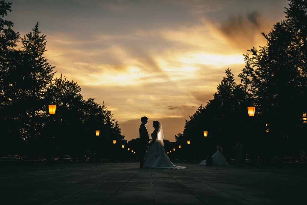 夕暮れの並木道で、街灯の明かりの中、向き合って見つめ合う新郎新婦のシルエット。