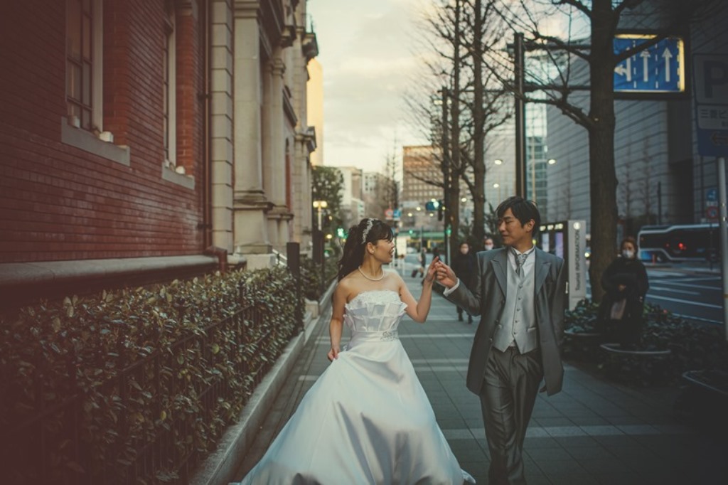 赤レンガの街並みを手をつなぎ歩く新郎新婦、街灯とビル群を背景に。