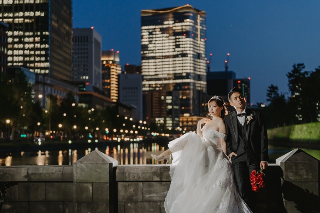 夜のビル群を背景に、赤いバラの花束を持ち石垣にもたれる新郎新婦。