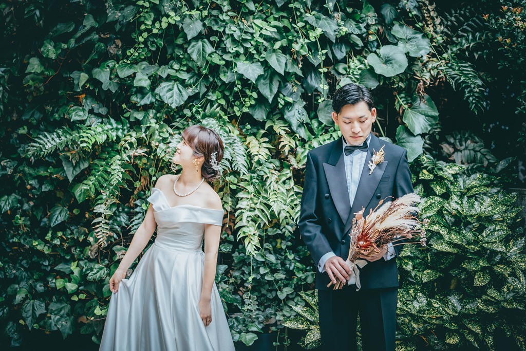 蔦の緑の壁を背景に、ドライブーケを持つ新郎とドレスの裾を持ち振り返る新婦。