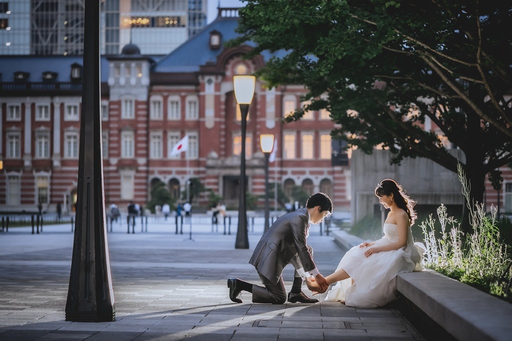 夕暮れの東京駅丸の内駅舎前、片膝をつき新婦の手を取る新郎、木々を背景に。