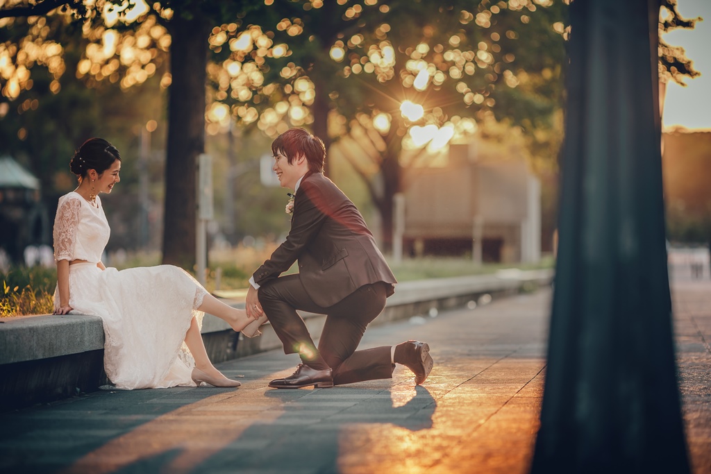 木漏れ日の輝く並木道で、片膝をつき新婦の靴を直す新郎、腰掛けて微笑む新婦。