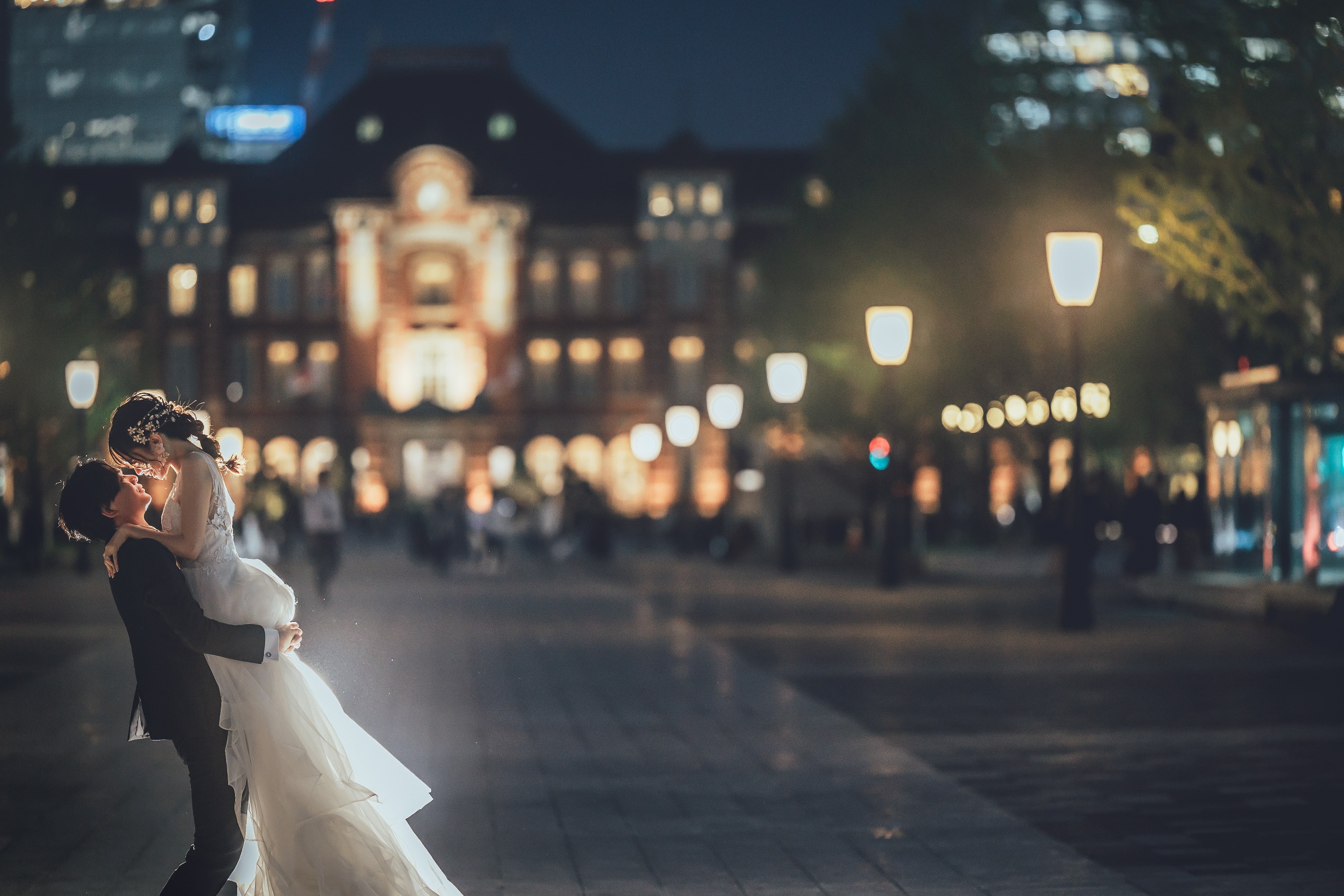 東京駅舎前の夜景を背景に、新郎が新婦を高く抱き上げる東京駅ならではのウェディングフォト。