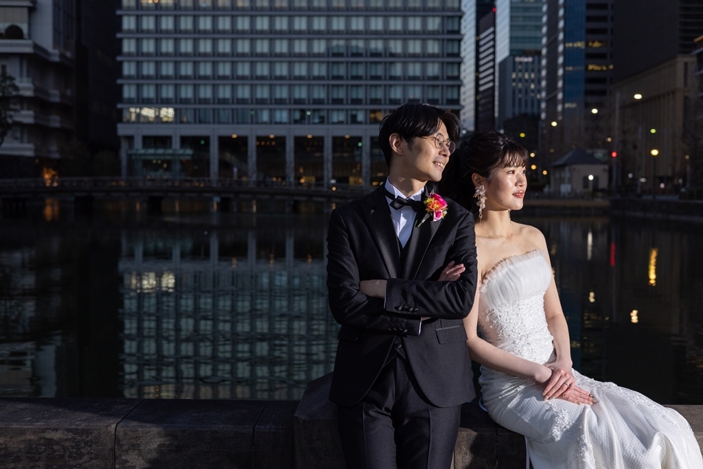 内堀端の石垣に腕を組んで並んで座り遠くを見つめる新郎新婦、夜のビル群を背景に。