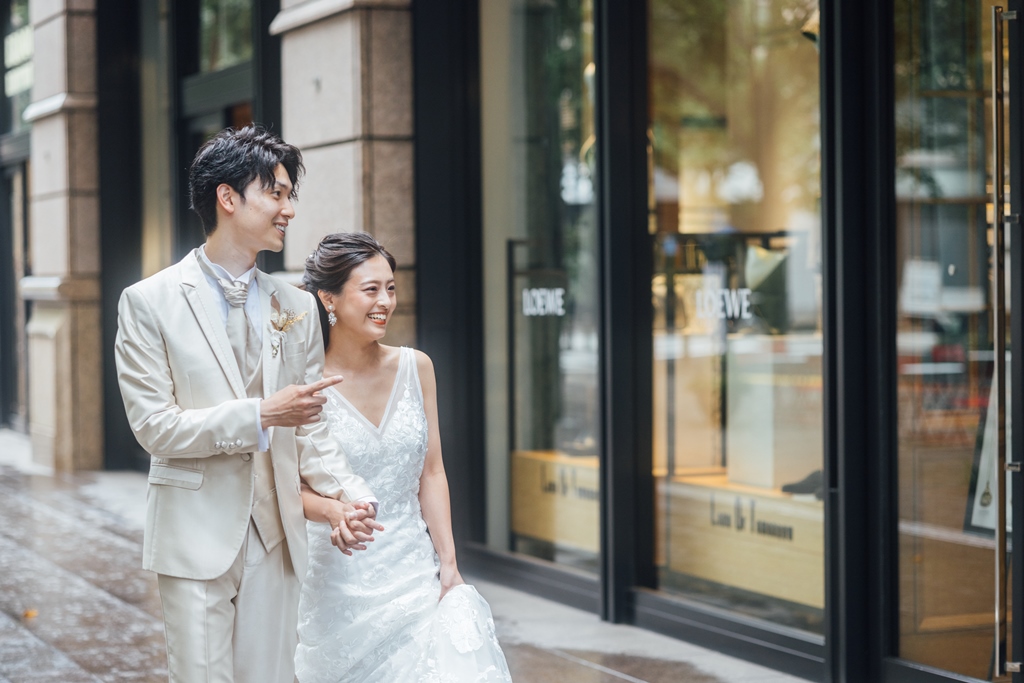 ハイブランドが並ぶ通りを、腕を組んで微笑みながら歩く新郎新婦、ベージュのスーツと白いドレス。