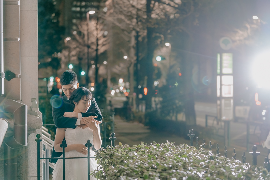 夜の街並みで、新婦の後ろから優しく抱きしめる新郎、植え込みと街灯を背景に。