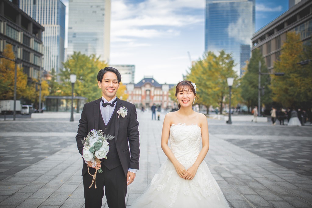 東京駅丸の内駅舎を背景に、笑顔でカメラを見つめる新郎新婦、白いブーケを手に。