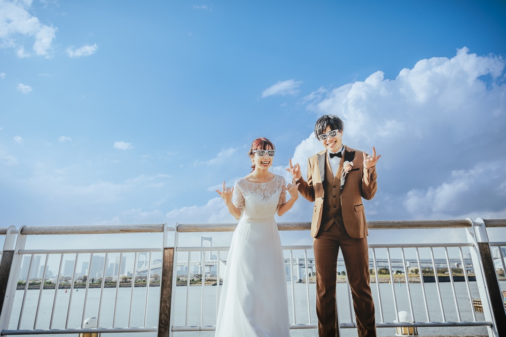 サングラス姿でピースサインをする楽しげな新郎新婦、お台場海浜公園の青空の下で。