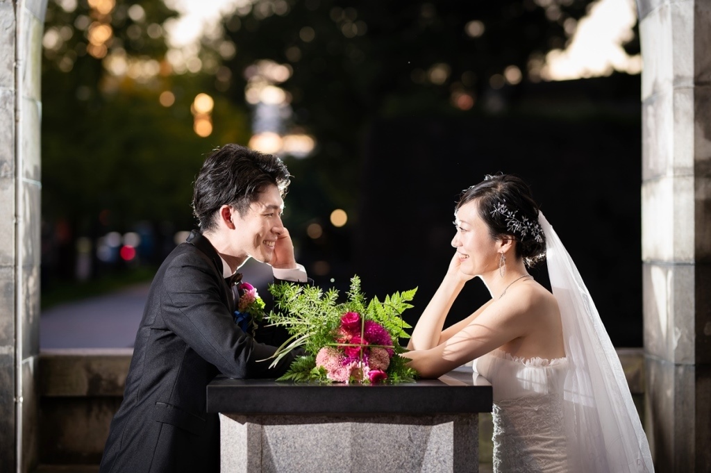 石造りテーブルに置かれたブーケ越しに、頬に手を添え微笑み合う新郎新婦、夕暮れの並木道を背景に。