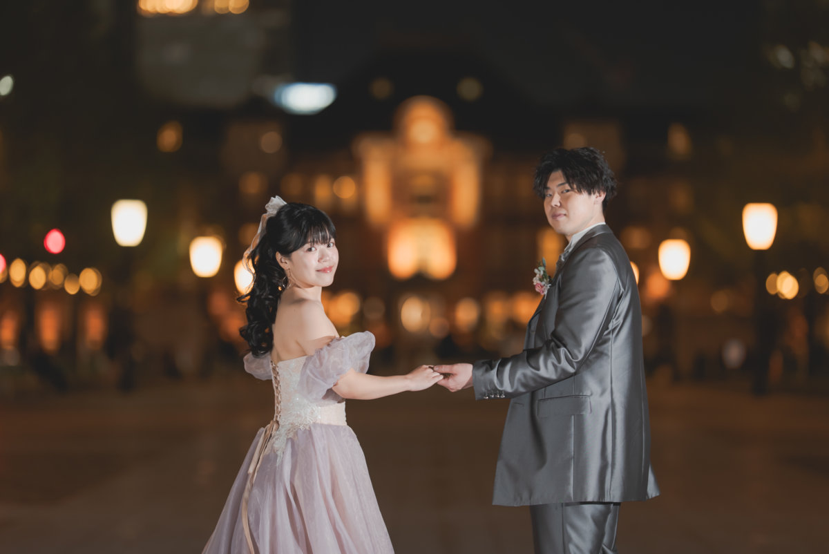 夜の東京駅前の広場で、手を取り合って向かい合う新郎とカラードレスの花嫁。駅舎の灯りをぼかした前撮り