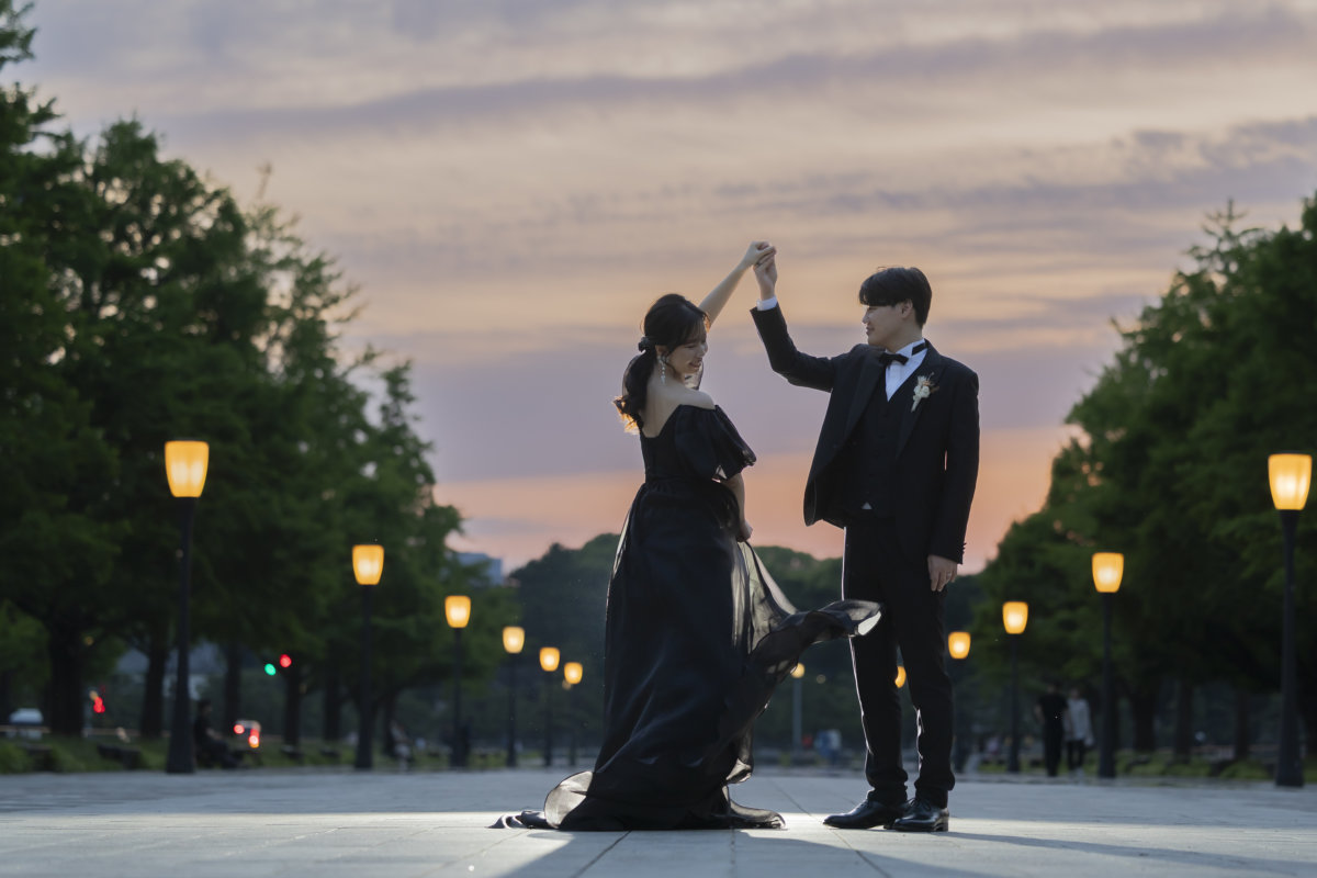夕暮れの行幸通りで、手を取り合ってダンスのようにくるりと回る新郎と黒いカラードレスの花嫁のウェディングフォト