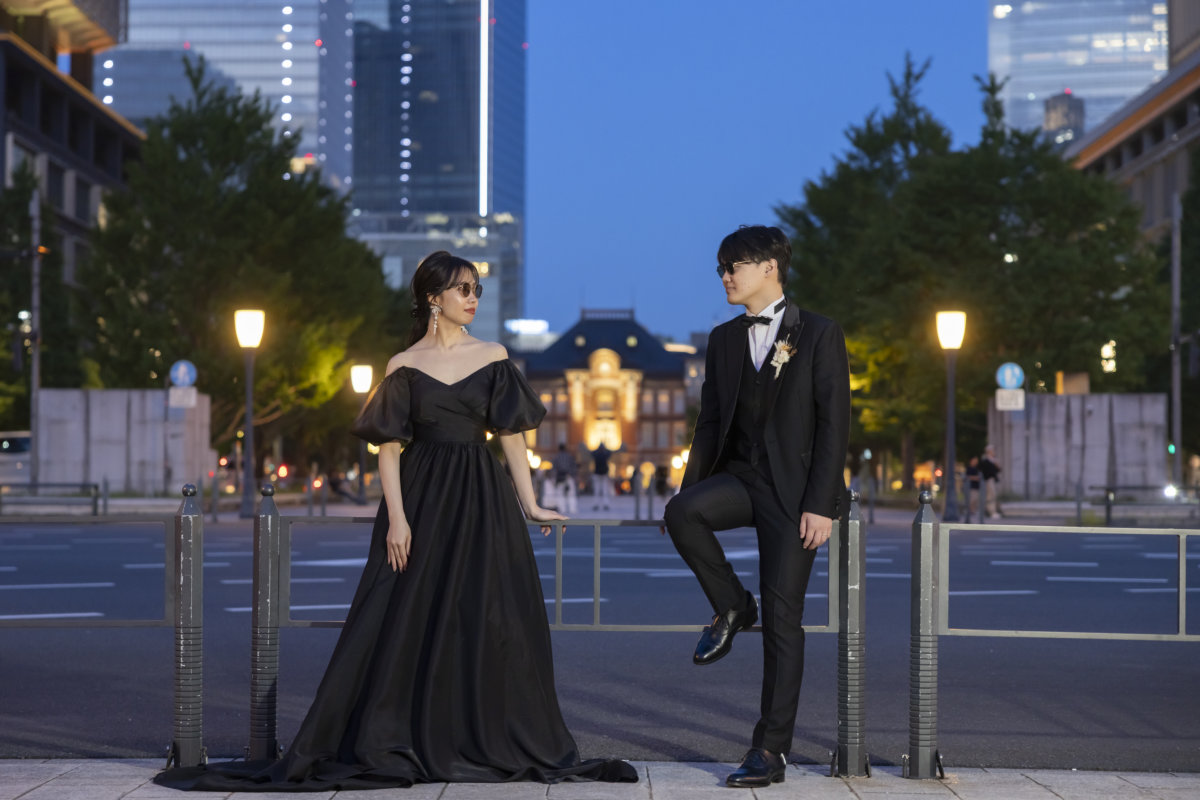 夜の東京駅丸の内駅舎を背に、柵に足をかけて並ぶサングラス姿の新郎と黒いカラードレスの花嫁の前撮り