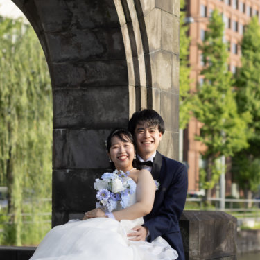 石造りのアーチ門の下、新郎に寄り添って笑顔を見せる新婦。ブルーのブーケとネイビーのタキシードが映える縦構図の前撮り