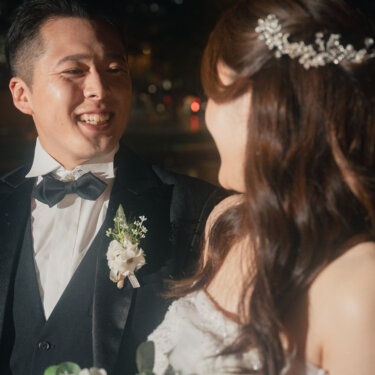 新婦を見つめて笑う黒タキシードの新郎のアップ。雨に濡れた東京駅前広場の灯りが背景にぼける夜の結婚写真