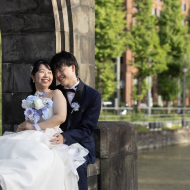 石造りのアーチ門のたもとに腰かけ、ブルーのブーケを抱えて笑う新婦と寄り添う新郎。柳と水辺、赤レンガのビルが背景の丸の内ロケーションフォト