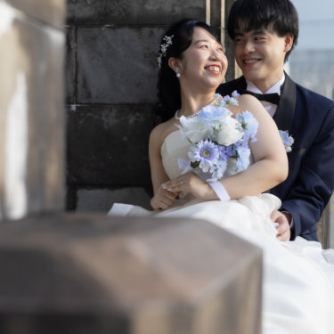 石造りの柱の陰に腰かけ、ブルーのブーケを抱えて笑い合う新郎新婦。ネイビーのタキシードと白いドレスのフォトウェディング