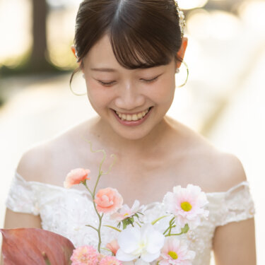 ピンクの花のブーケを胸元に抱え、うつむいてほほえむ花嫁のアップ。やわらかな光に包まれたウェディングフォト