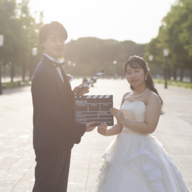 夕方の並木道でカチンコを持って笑うネイビーのタキシードの新郎とウェディングドレスの花嫁。東京駅に近い丸の内での前撮りのカチンコカット