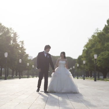 街灯が並ぶ広い並木道を手をつないで歩く新郎新婦。東京駅前の通りで撮る洋装フォトウェディング