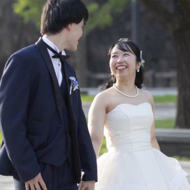 石垣と緑を背に見つめ合って笑うネイビーのタキシードの新郎とパールのネックレスの花嫁の縦位置の一枚。丸の内の石垣沿いで撮る洋装前撮り