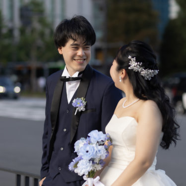 街の歩道に腰かけて見つめ合う新郎新婦。ブルーのブーケとネイビータキシードで撮る丸の内の洋装前撮り