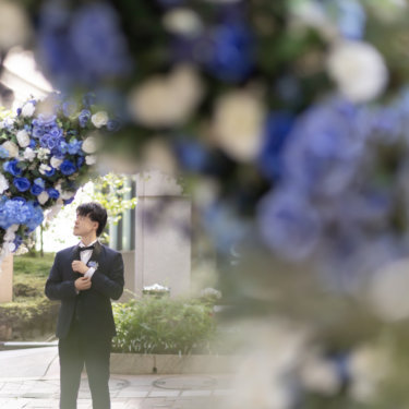 青と白の花のアーチ越しに佇む新郎。ネイビータキシードで撮る街のロケーションフォト