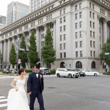 石造りのビルと高層ビルを見上げる構図で、横断歩道を渡る新郎新婦を写した東京駅周辺の街ロケ前撮り