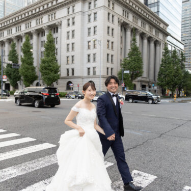 円柱が並ぶクラシカルなビルを背に、ドレスの裾を持って横断歩道を歩く花嫁と新郎。丸の内の街並みで撮るフォトウェディング
